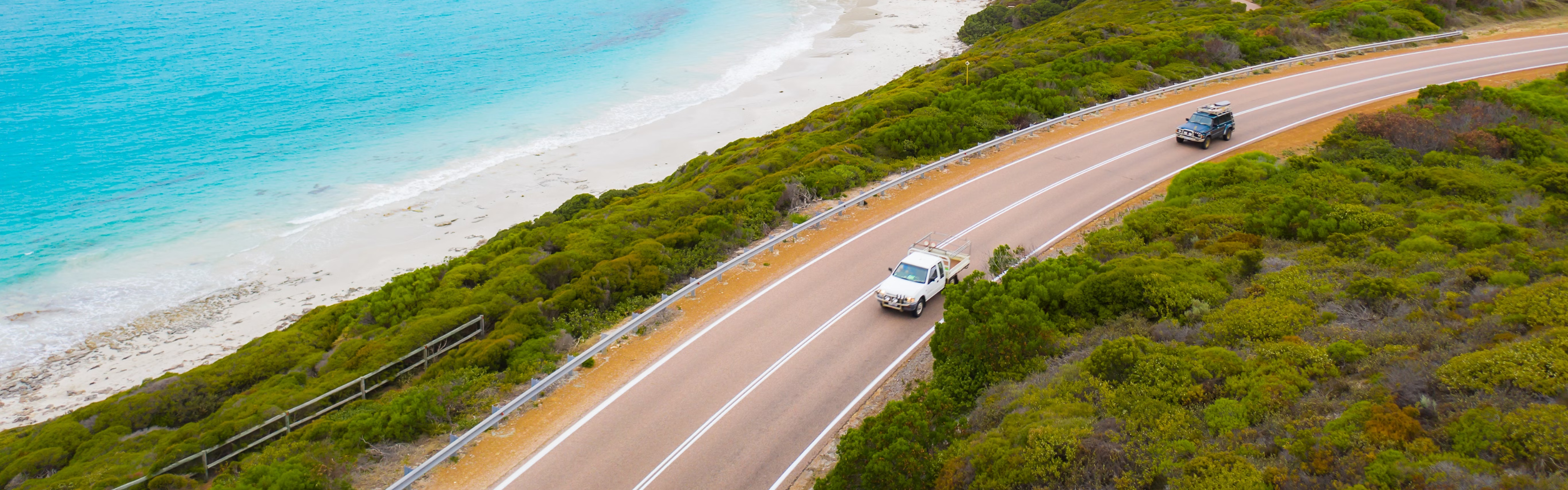 Two cars driving on a scenic coastal road with turquoise water and white sand.
